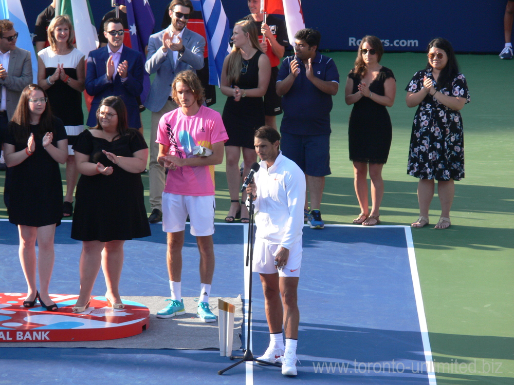 Rafael Nadal in front of microphone, his Trophy and Stefanos Tsitsipas on the left, also holding his Trophy.